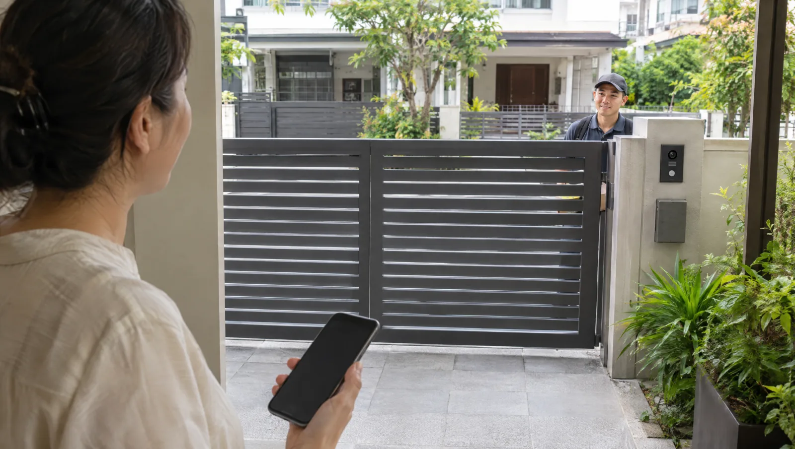 Homeowner holding a phone with a driveway gate and gatepost intercom in view.