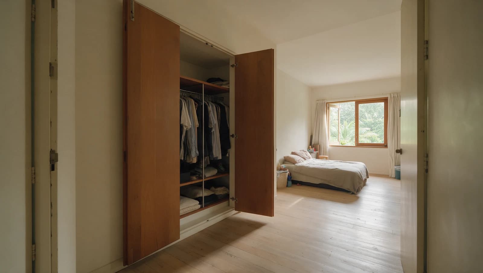 Two hinged wardrobe doors standing wide open into a bedroom, with clear floor in front of the wardrobe and the bed further back by the window.